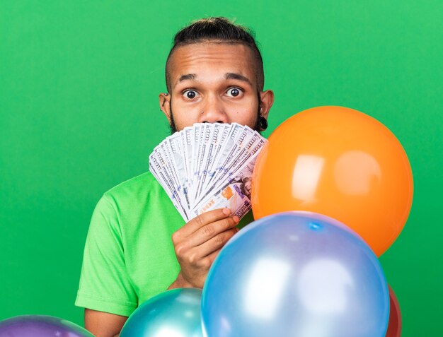 Chico afroamericano joven sorprendido con camiseta verde de pie detrás de globos con la cara cubierta con dinero en efectivo aislado en la pared verde