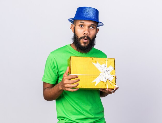 Chico afroamericano joven sorprendido con sombrero de fiesta con caja de regalo aislada en la pared blanca