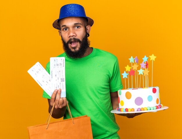 Chico afroamericano joven sorprendido con sombrero de fiesta sosteniendo una bolsa de regalo y pastel con boletos aislados en la pared naranja