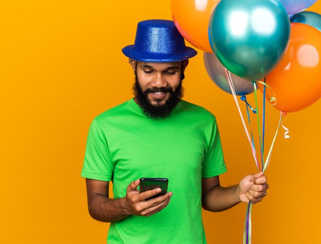 Chico afroamericano joven sorprendido con sombrero de fiesta sosteniendo y mirando el teléfono aislado en la pared naranja