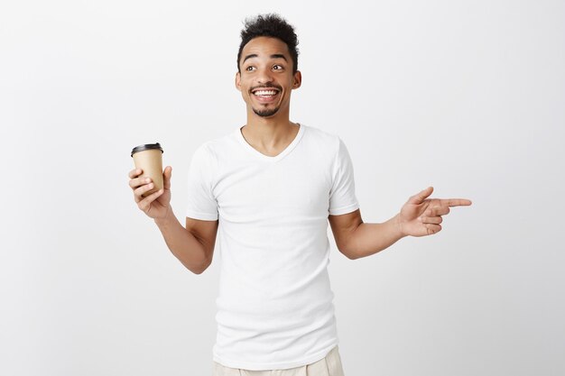 Chico afroamericano sonriente alegre en camiseta blanca apuntando a la derecha, sosteniendo la taza con café