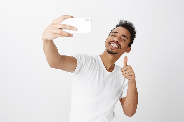 Chico afroamericano sonriente descarado tomando selfie en smartphone, mostrando gesto de pulgar hacia arriba