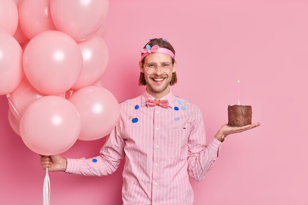 Chico alegre sin afeitar con peinado de moda se prepara para la celebración de cumpleaños sostiene pastel de chocolate y globos de helio se divierte en la fiesta recibe felicitaciones de amigos confeti volando alrededor