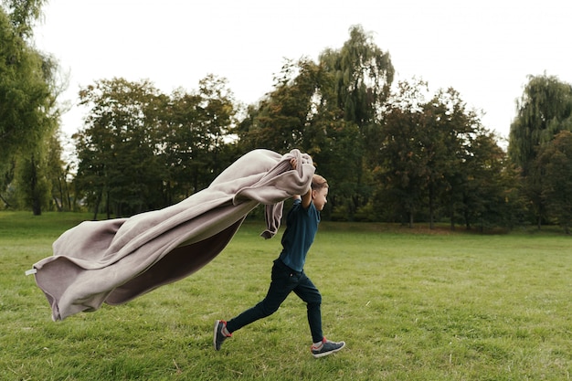 Foto gratuita chico alegre corriendo con una manta voladora en el parque