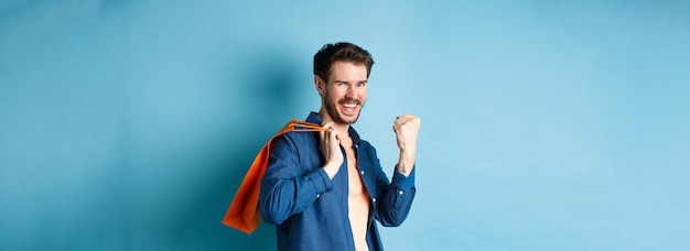 Foto gratuita chico alegre diciendo sí levantando el puño y sonriendo sosteniendo una bolsa de compras naranja sintiendo alegría después