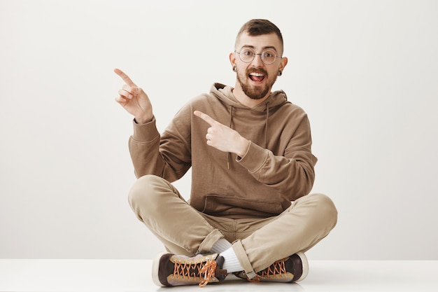 Foto gratuita chico alegre guapo que señala el dedo a la izquierda y sentarse en el suelo