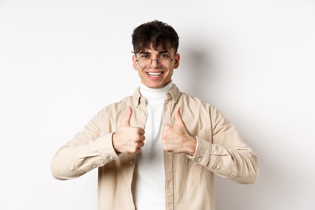 Chico alegre hipster con gafas mostrando los pulgares hacia arriba y sonriendo, como buen producto, recomendar o elogiar el anuncio, de pie sobre fondo blanco.