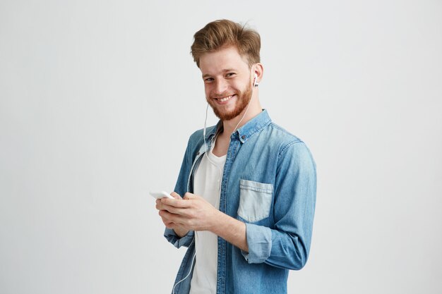 Chico alegre joven en auriculares sonriendo sosteniendo teléfono escuchando música.