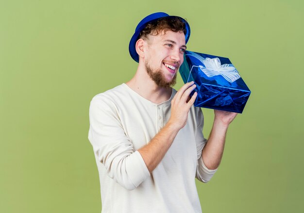 Chico alegre joven guapo partido eslavo con sombrero de fiesta mirando recto sosteniendo la caja de regalo aislada sobre fondo verde oliva con espacio de copia