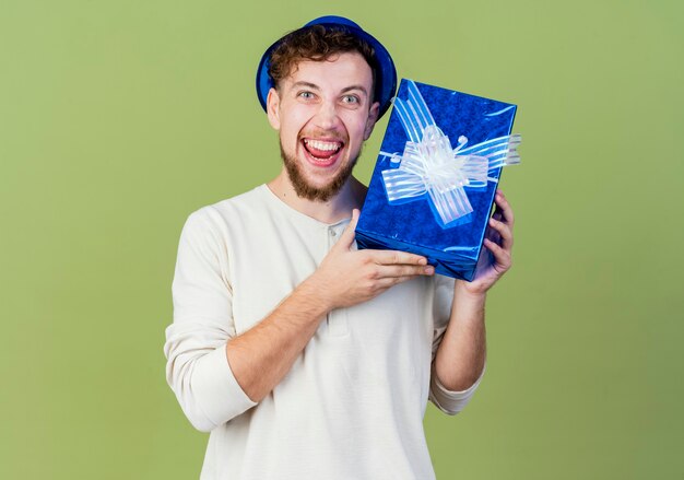 Chico alegre joven guapo partido eslavo con sombrero de fiesta mostrando caja de regalo mirando a cámara aislada sobre fondo verde oliva con espacio de copia