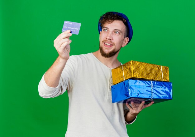 Chico alegre joven guapo partido eslavo con sombrero de fiesta sosteniendo cajas de regalo y tarjeta de crédito mirando la tarjeta aislada en la pared verde