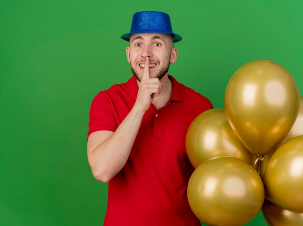 Chico alegre joven guapo partido eslavo con sombrero de fiesta sosteniendo globos mirando a la cámara haciendo gesto de silencio aislado sobre fondo verde con espacio de copia