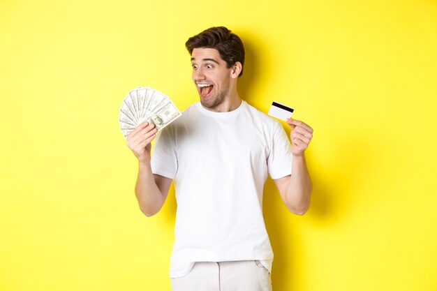 Chico alegre mirando dinero, sosteniendo tarjeta de crédito, concepto de crédito bancario y préstamos, de pie sobre fondo amarillo