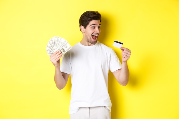 Chico alegre mirando tarjeta de crédito, sosteniendo dinero, concepto de crédito bancario y préstamos, de pie sobre fondo amarillo
