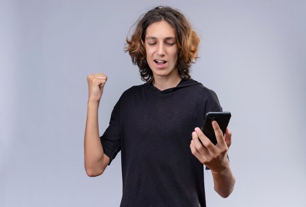 Chico alegre con pelo largo en camiseta negra sosteniendo un teléfono en la pared blanca