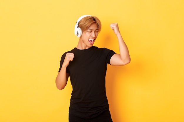 Chico asiático atractivo despreocupado con cabello rubio, cantando y bailando mientras escucha música en auriculares inalámbricos, de pie pared amarilla