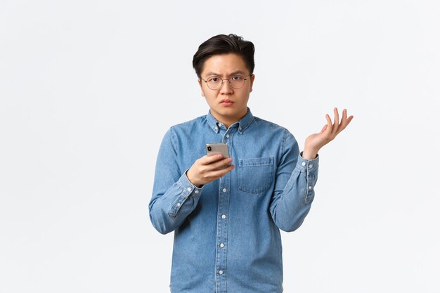 Chico asiático confundido y decepcionado con gafas no puede entender las razones, de pie fondo blanco, levantando la mano desconcertado después de ver algo frustrante en el teléfono móvil, fondo blanco.