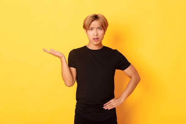 Chico asiático frustrado y molesto con cabello rubio, levantando la mano confundida, pared amarilla