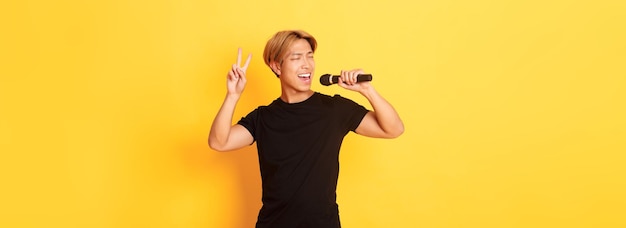 Chico asiático guapo despreocupado interpretando canciones cantando en el micrófono y mostrando gesto de paz standi