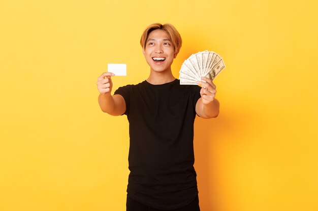 Chico asiático guapo feliz que mira la esquina superior izquierda pensativa y complacida mientras muestra dinero y tarjeta de crédito, pared amarilla