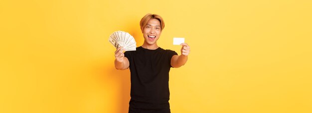 Foto gratuita chico asiático guapo feliz en ropa casual negra mostrando dinero y tarjeta de crédito sonriendo grito descarado