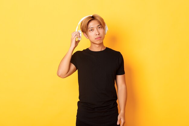 Chico asiático guapo joven con cabello rubio, auriculares de despegue para escucharte, pared amarilla de pie