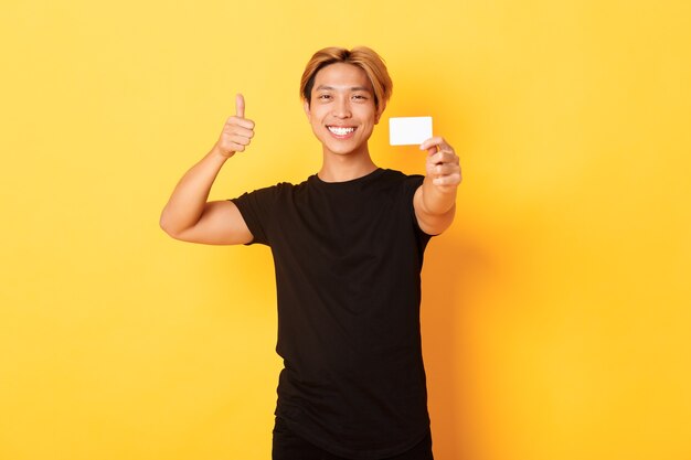 Chico asiático guapo satisfecho sonriendo complacido y mostrando tarjeta de crédito, pulgar hacia arriba en aprobación, pared amarilla de pie