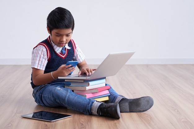 Chico asiático sentado en el piso con tableta y computadora portátil en libros apilados y con smartphone