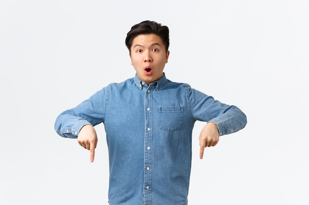 Un chico asiático sorprendido e impresionado con camisa azul, apuntando con el dedo hacia abajo y mirando a la cámara sin palabras, haciendo preguntas sobre el producto, encontró algo interesante, de pie con un fondo blanco.