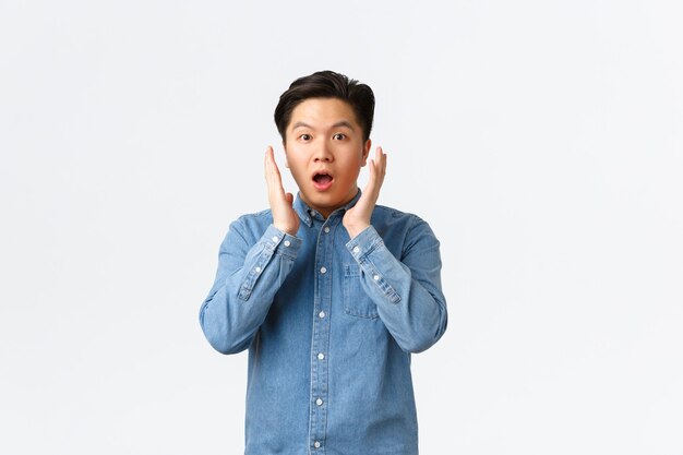 Un chico asiático sorprendido e impresionado reacciona al gran anuncio, tomados de la mano cerca de la cara y jadeando, mirando algo increíble, de pie asombrado sobre el fondo blanco.