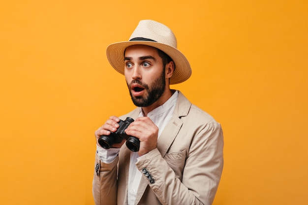 Chico asombrado con sombrero sosteniendo binoculares en pared naranja