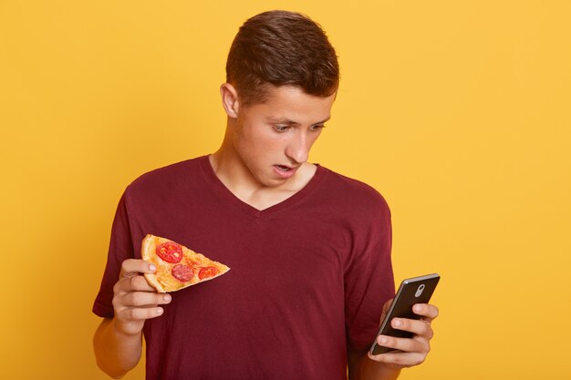 Un chico asombroso y guapo que sostiene una rebanada de sabrosa pizza y teléfono, revisa la red social mientras come algo, ve noticias de frenado