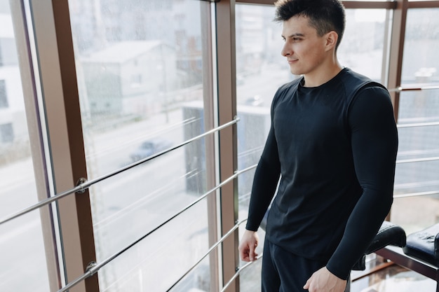 Chico atractivo deportivo junto a la ventana. Atleta posando cerca de amplias ventanas. Gimnasio y deportes.