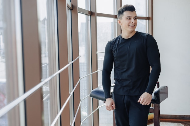 Chico atractivo deportivo junto a la ventana. Atleta posando cerca de amplias ventanas. Gimnasio y deportes.