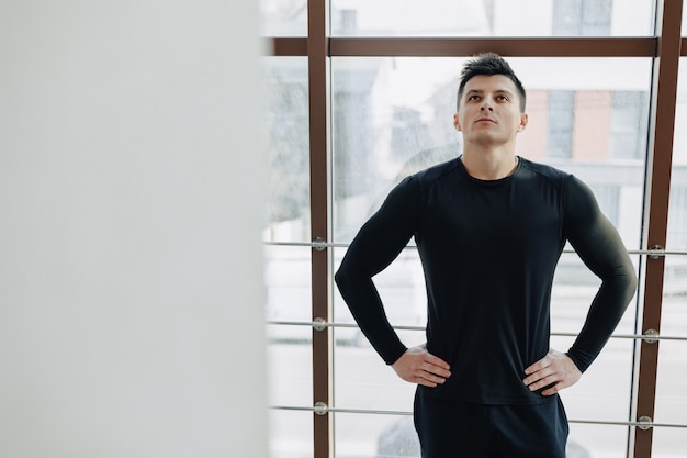 Chico atractivo deportivo junto a la ventana. Atleta posando cerca de amplias ventanas. Gimnasio y deportes.