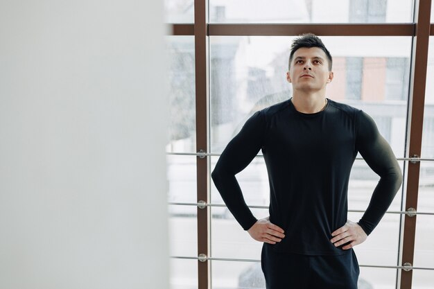 Chico atractivo deportivo junto a la ventana. Atleta posando cerca de amplias ventanas. Gimnasio y deportes.