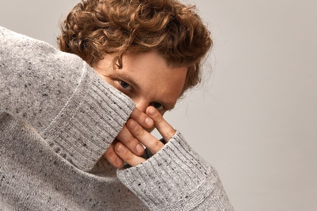 Chico atractivo hipster con cabello pelirrojo ondulado cruzando los dedos sobre su rostro con expresión facial penetrante, vestido con jersey gris. Copyspace para tu información