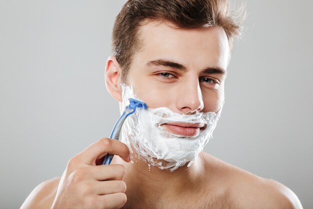 Chico atractivo con el pelo corto y oscuro afeitándose la cara con una maquinilla de afeitar y gel o crema aislado sobre la pared gris de cerca