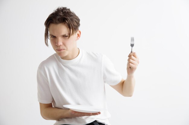 Chico atractivo triste joven con plato vacío y tenedor aislado en la pared gris.