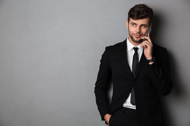 Chico barbudo alegre en traje negro hablando por teléfono móvil, mirando a un lado