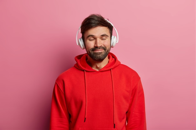 Un chico barbudo complacido disfruta escuchando música en auriculares estéreo, cierra los ojos y sonríe suavemente, usa una sudadera roja, se siente bien, modela sobre una pared rosa pastel. Adolescentes, hobby, concepto de estilo de vida