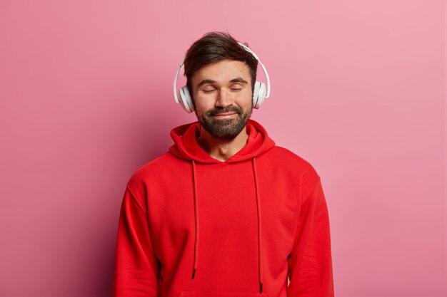 Un chico barbudo complacido disfruta escuchando música en auriculares estéreo, cierra los ojos y sonríe suavemente, usa una sudadera roja, se siente bien, modela sobre una pared rosa pastel. Adolescentes, hobby, concepto de estilo de vida