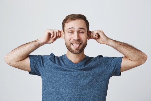 Chico barbudo despreocupado divertido que tira de las orejas y muestra la lengua tonta