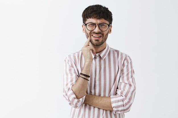 Chico barbudo elegante confundido posando contra la pared blanca con gafas