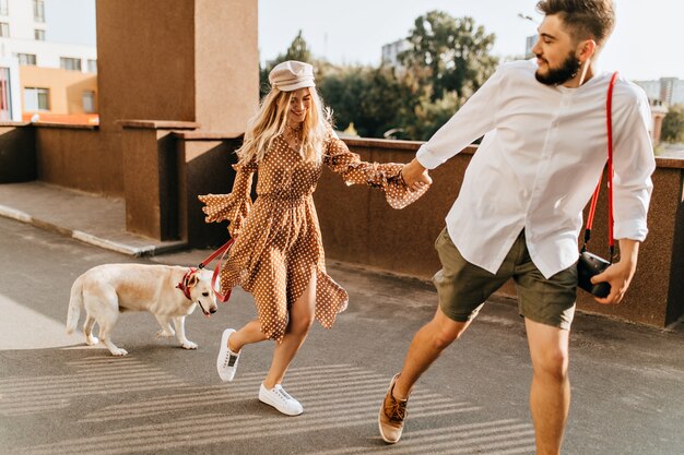 Chico barbudo en pantalones cortos de color caqui sostiene a la chica rubia con la mano y corre. Pareja disfrutando de un paseo de verano con perro.