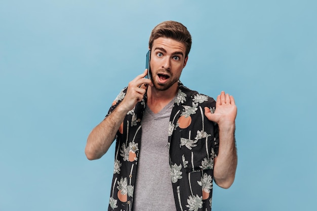 Chico barbudo sorprendido con ojos azules en camisa de verano moderna habla por teléfono y mira sorprendido a la cámara en un contexto aislado