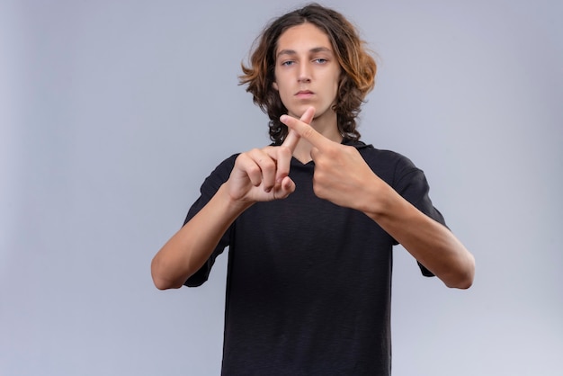 Chico con cabello largo en camiseta negra cruzó los dedos en la pared blanca