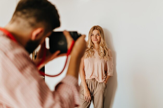 Chico de cabello oscuro hace una foto de su novia rubia vestida con una camiseta rosa y pantalones beige contra una pared blanca