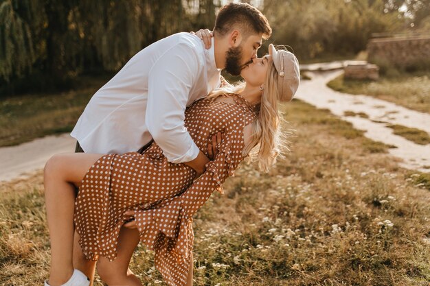 Chico con camisa blanca inclinando a su novia durante un beso suave y apasionado. Pareja abrazada en medio del parque.