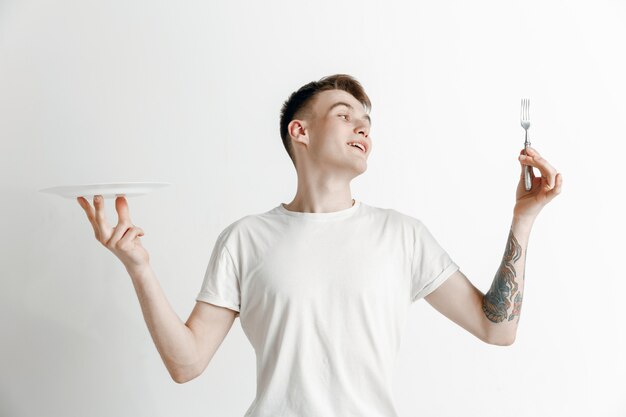 Chico caucásico atractivo sonriente joven con plato vacío y tenedor aislado sobre fondo gris. Copie el espacio y simule. Fondo de plantilla en blanco.